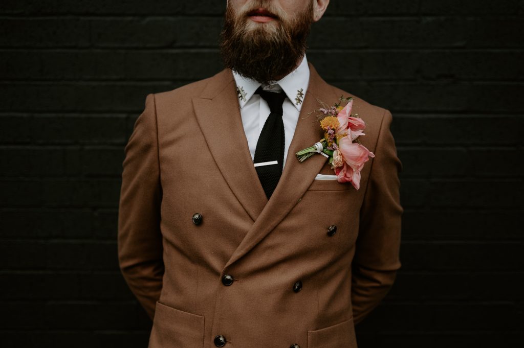 Details of a grooms tobacco brown suit and collar pins whilst stood against a black wall at Five Four Studios.