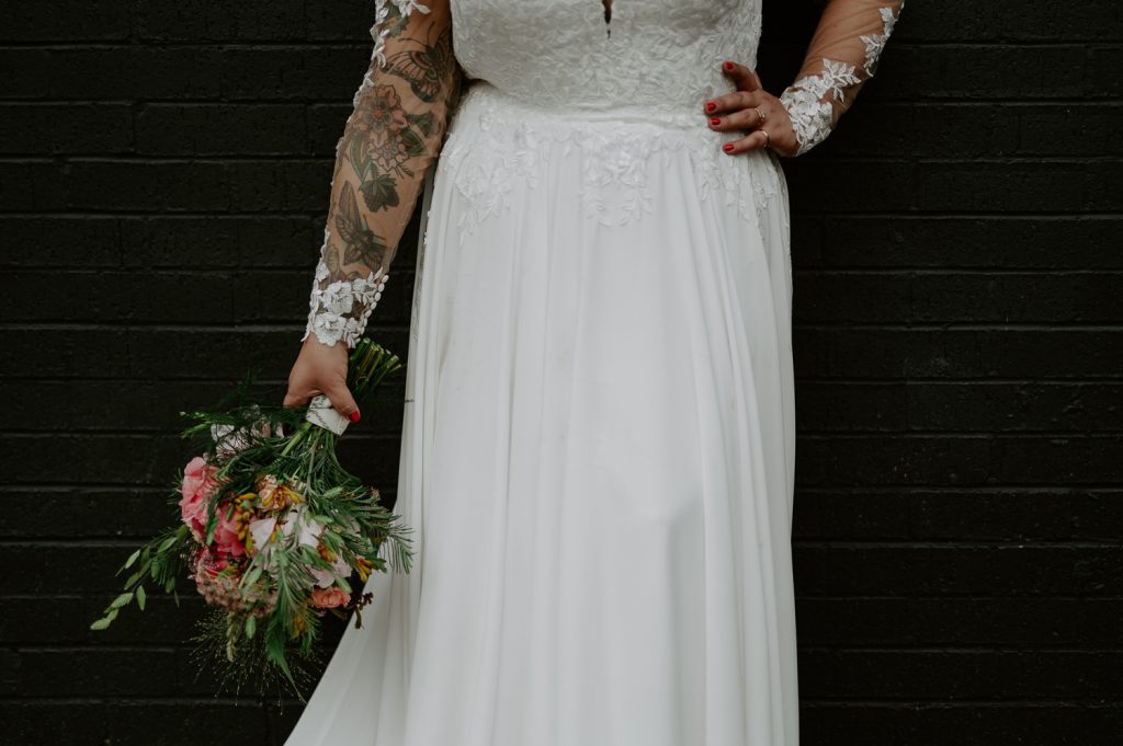 Details of a brides dress and tattoos whilst stood against a black wall at Five Four Studios.