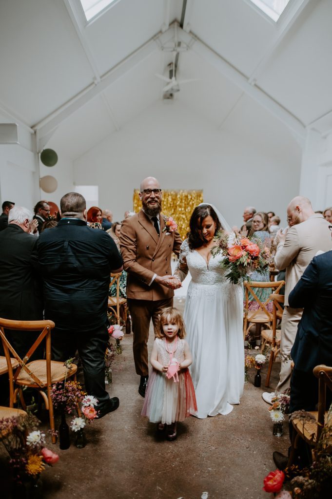 A bride, groom and their daughter walk up the aisle at their stylishly colourful wedding at Five Four Studios in Manchester.