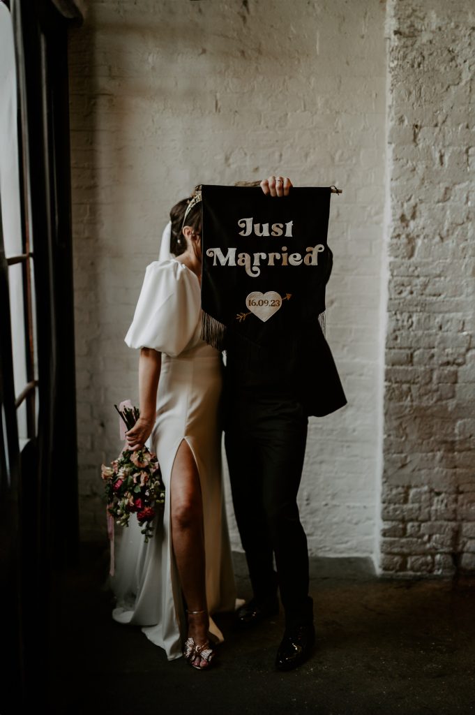 A bride and groom hold up a black JUST MARRIED custom wedding banner at 100 Barrington.
