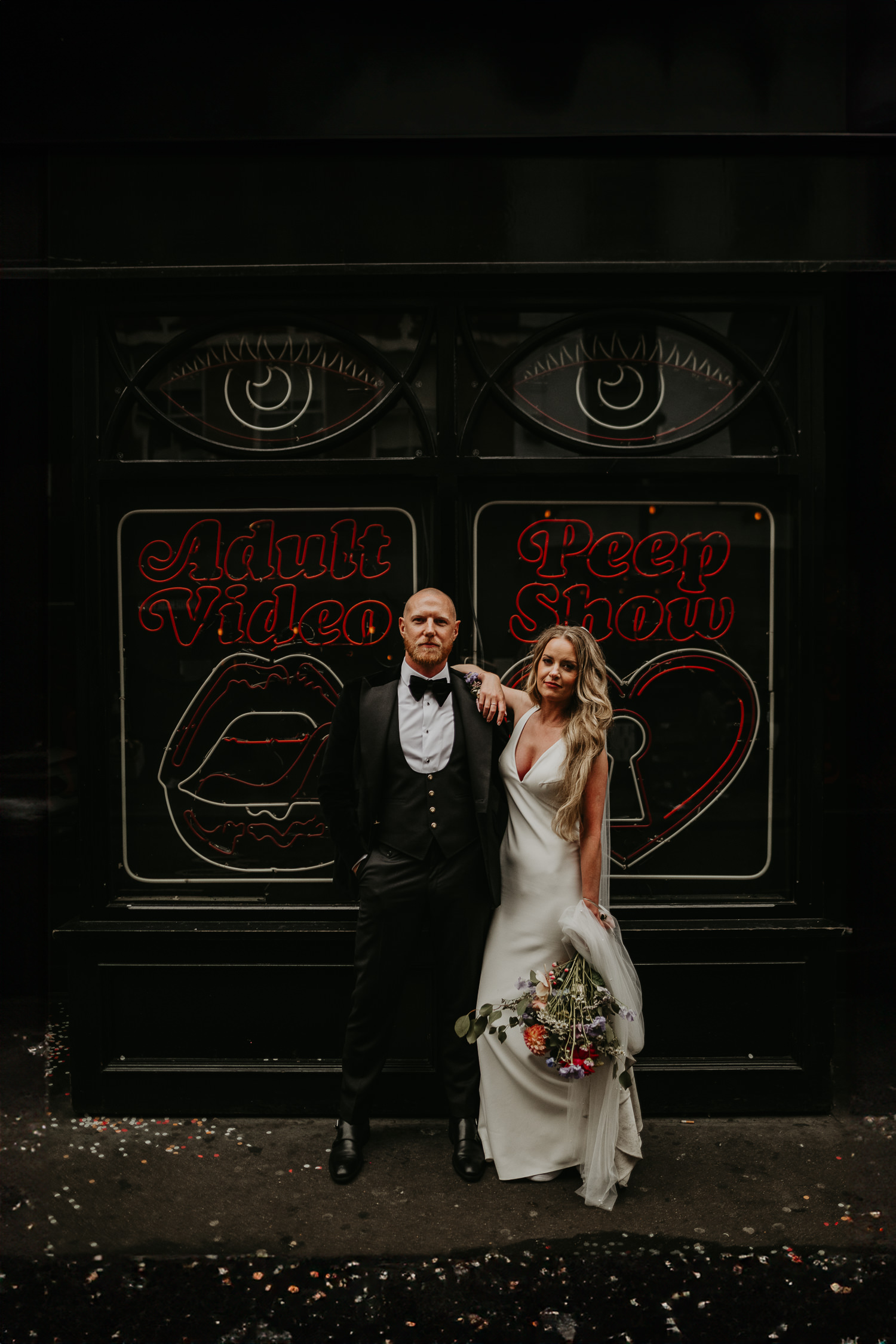 A bride and groom pose in front of a peep show neon in London.