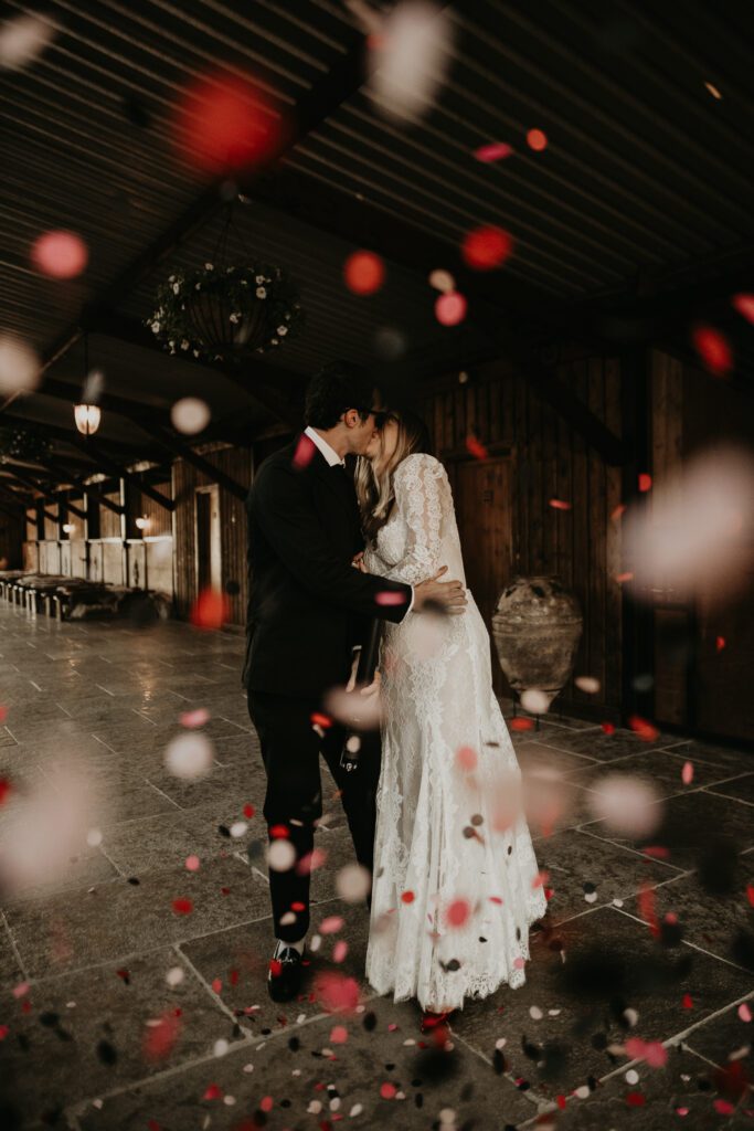 Bride and groom kiss whilst pink and red confetti fall on them inside the stables at The Willow Marsh Farm. 