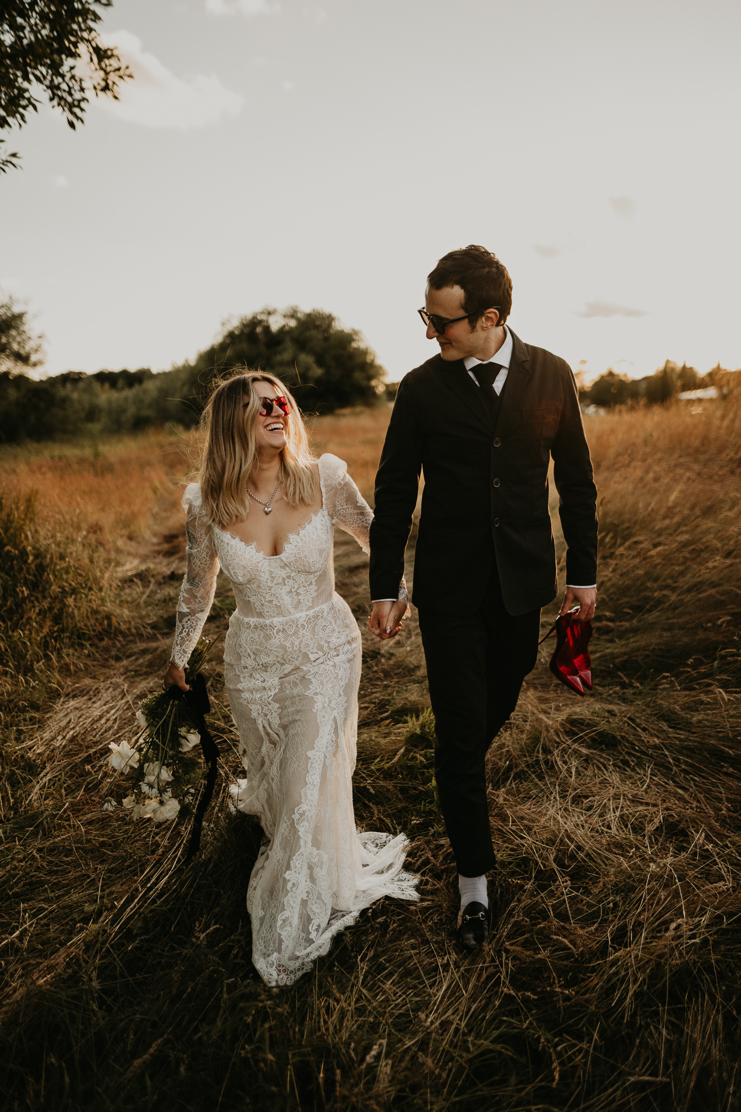 A bride and groom walk through the long grass at golden hour as the groom holds his bride's red Christian louboutin shoes.