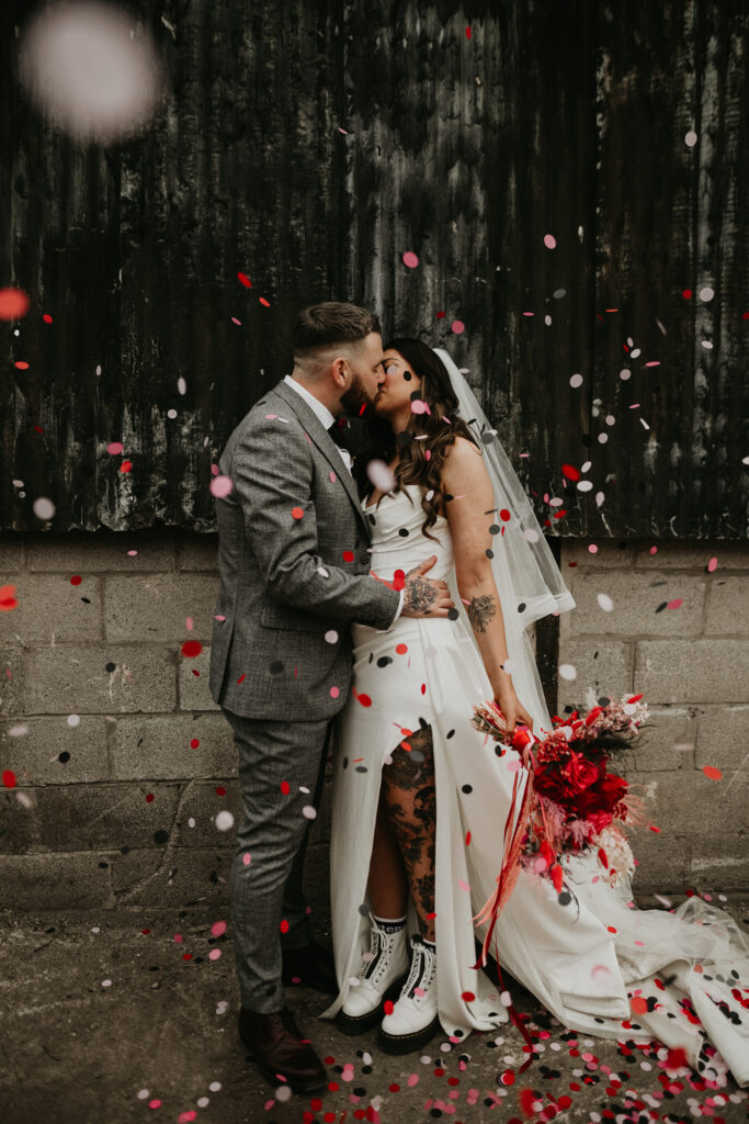 A tattooed bride wearing Dr Marten boots kissing her groom as biodegradable wedding confetti falls on them.