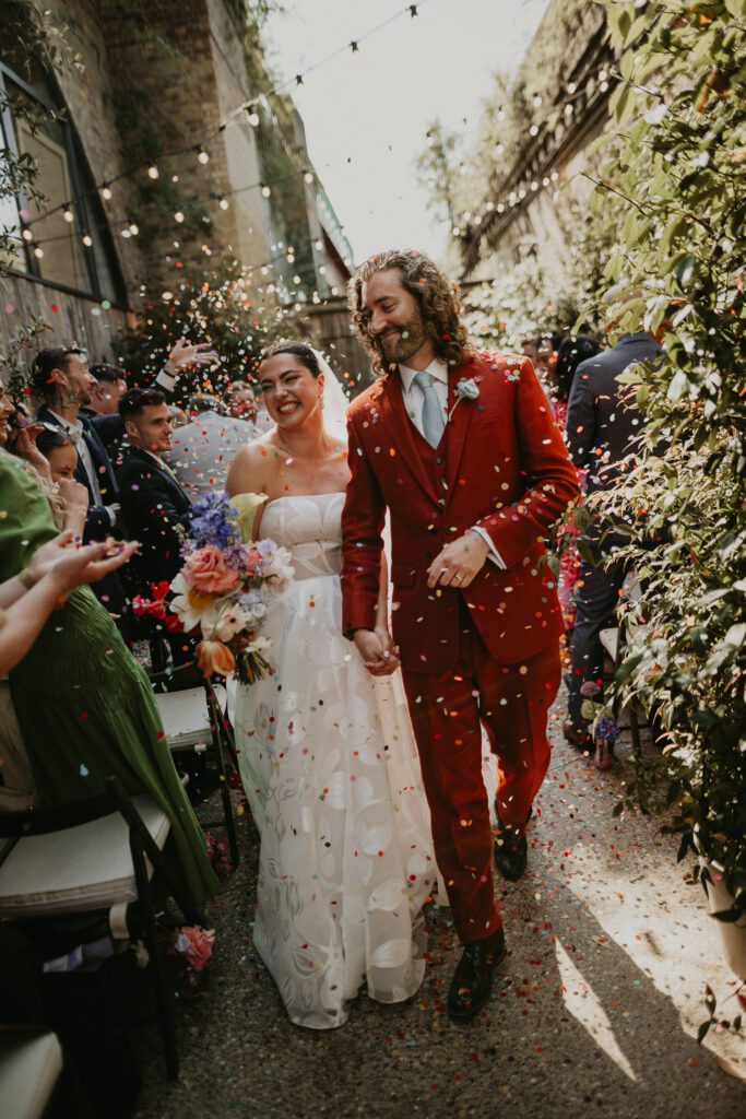 A bride and groom walk up the aisle at their wedding at 100 Barrington as their guests throw biodegradable wedding confetti.