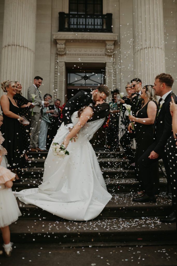 Bride and groom do the top kiss on the stairs at The Old Marylebone Town Hall whilst their guests throw biodegradable wedding confetti.